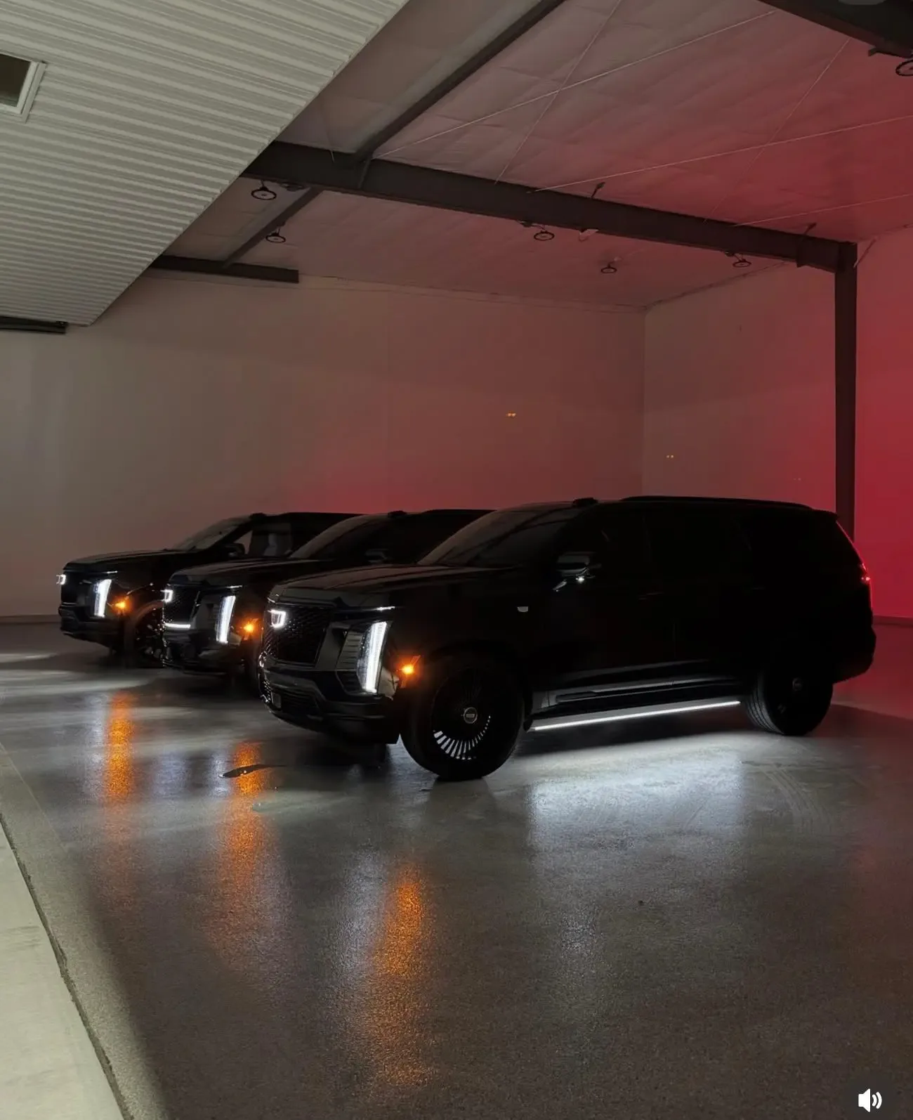 Three Cadillac Escalades with lit headlights in a dark garage