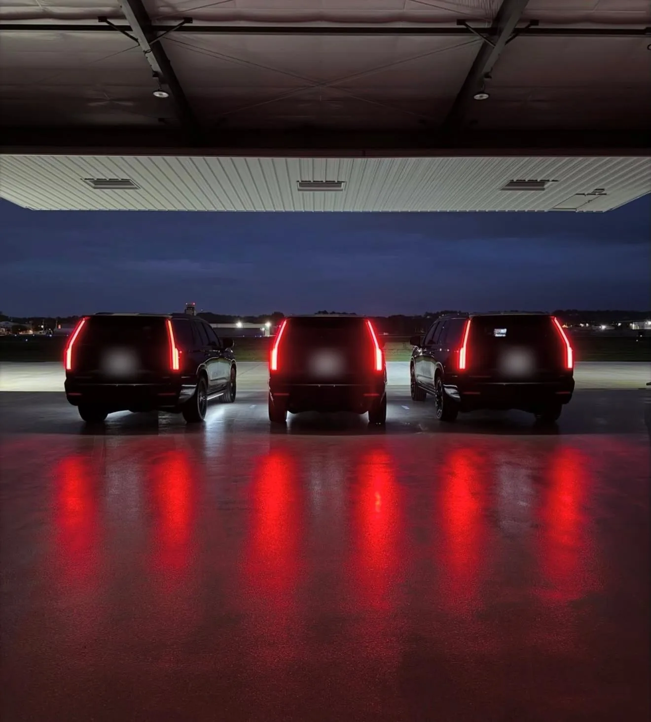 Three Cadillac Escalades from behind, tail lights reflecting on a hangar floor